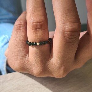 May include: A beaded ring with small, dark green beads and gold-coloured beads. The ring is worn on a finger, highlighting its delicate design. The ring is a piece of jewellery.