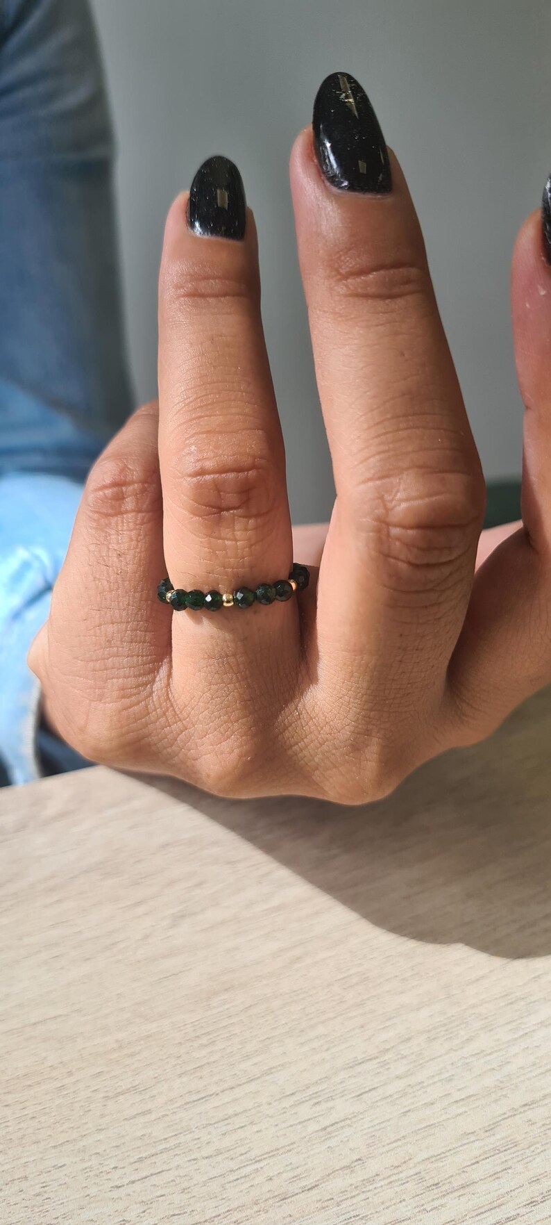 May include: A beaded ring with small, dark green beads and gold-coloured beads. The ring is worn on a finger, highlighting its delicate design. The ring is a piece of jewellery.
