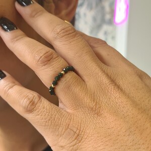 May include: A beaded ring featuring dark green gemstones and small gold beads. The ring is worn on a finger with black nail polish. The ring is a piece of jewellery.