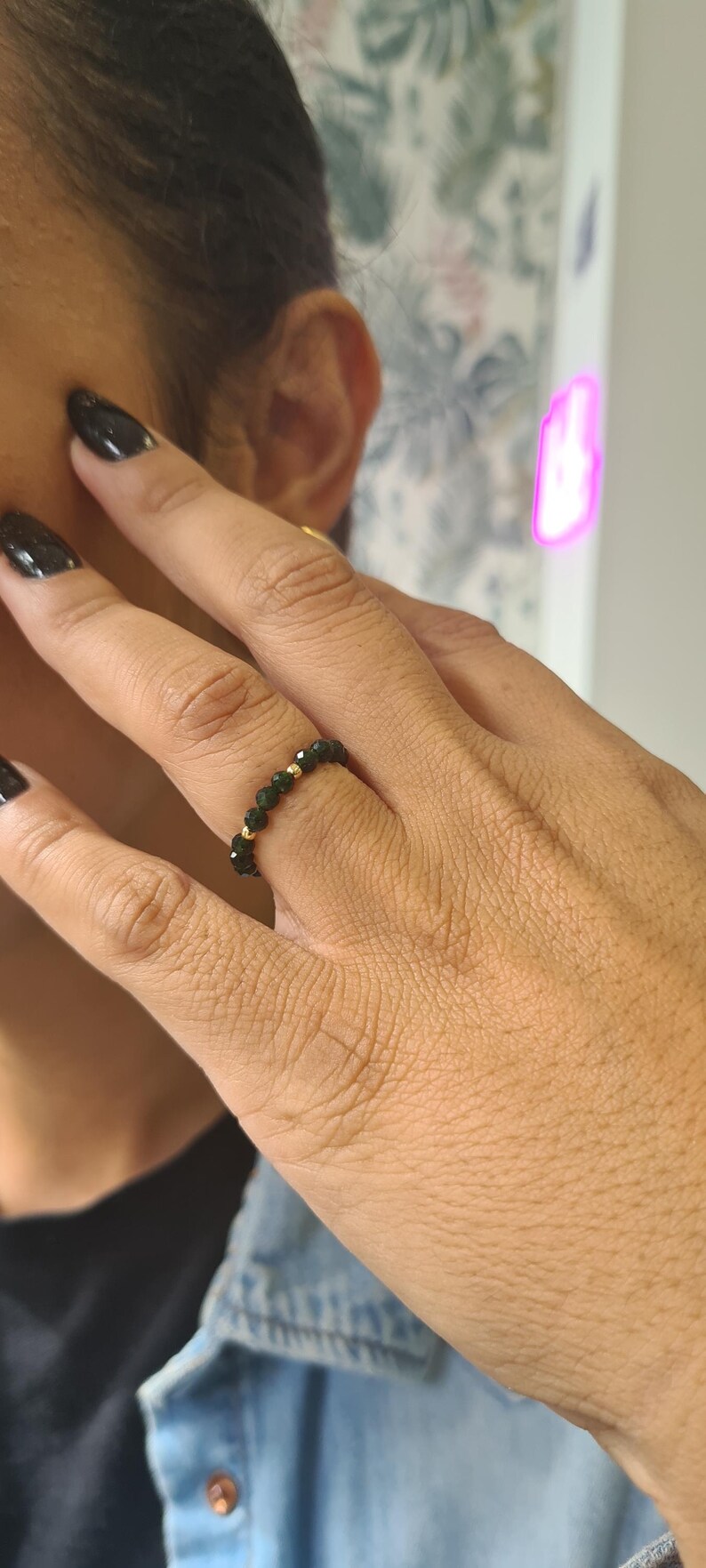 May include: A beaded ring featuring dark green gemstones and small gold beads. The ring is worn on a finger with black nail polish. The ring is a piece of jewellery.