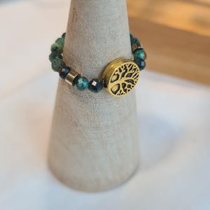 May include: A beaded ring with green and gold beads and a gold tree of life charm. The ring is displayed on a light-colored, cone-shaped stand. The ring is a piece of jewelry.