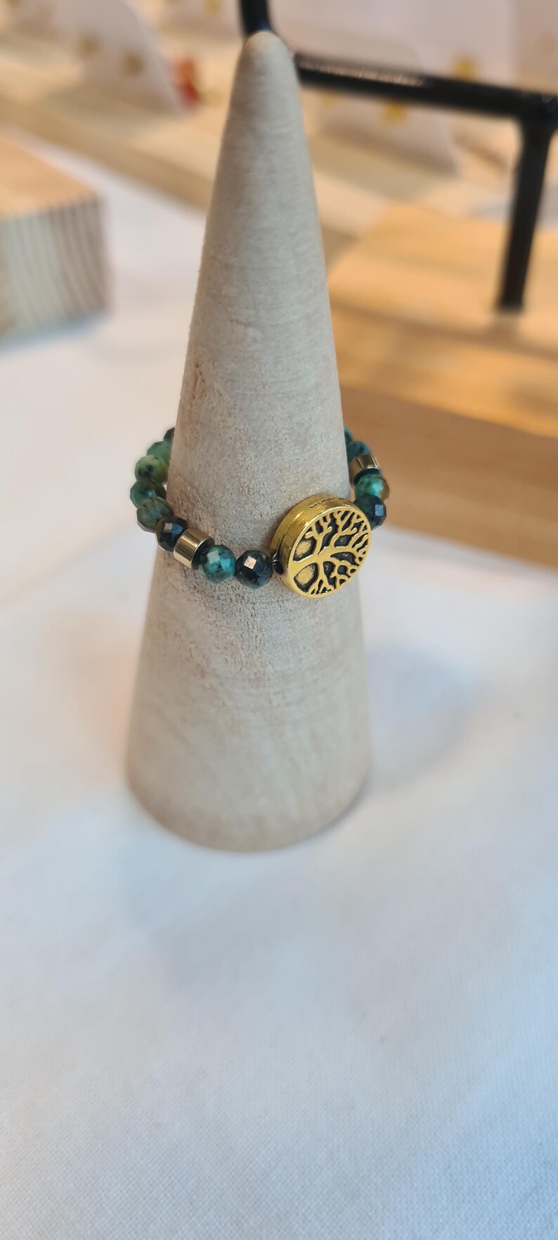May include: A beaded ring with green and gold beads and a gold tree of life charm. The ring is displayed on a light-colored, cone-shaped stand. The ring is a piece of jewelry.