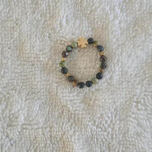 Puede incluir: Un anillo de cuentas con un dije de tr&eacute;bol de cuatro hojas de color dorado. El anillo est&aacute; hecho de peque&ntilde;as cuentas redondas en tonos verdes, marrones y negros, con espaciadores dorados. El anillo se muestra sobre una superficie blanca texturizada.