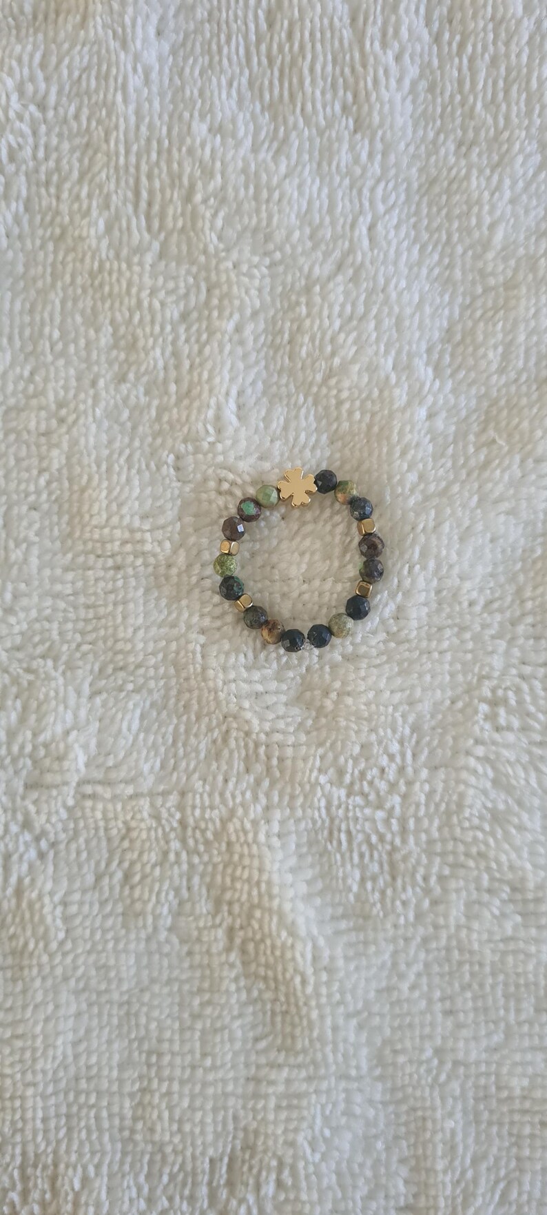 Puede incluir: Un anillo de cuentas con un dije de tr&eacute;bol de cuatro hojas de color dorado. El anillo est&aacute; hecho de peque&ntilde;as cuentas redondas en tonos verdes, marrones y negros, con espaciadores dorados. El anillo se muestra sobre una superficie blanca texturizada.