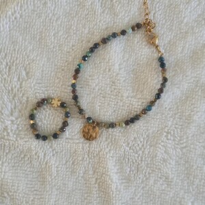 Puede incluir: Un conjunto de pulsera y anillo de cuentas. La joyer&iacute;a presenta peque&ntilde;as cuentas redondas en tonos verdes, azules y dorados. La pulsera tiene una cadena dorada y un dije dorado martillado. El anillo tiene un engaste dorado.