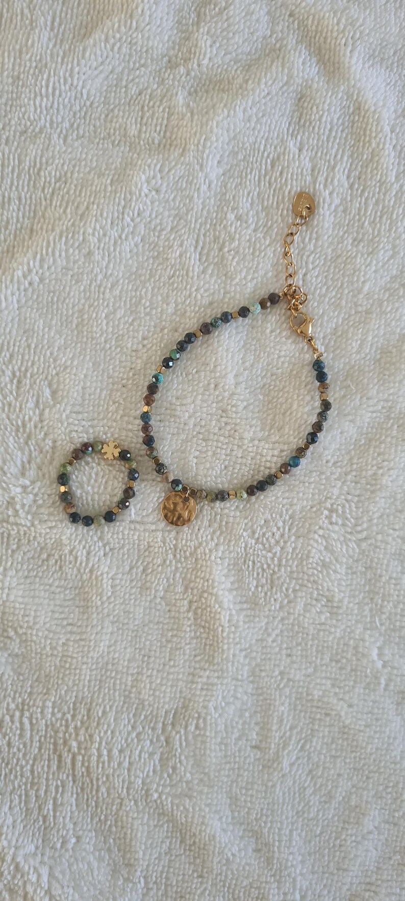 Puede incluir: Un conjunto de pulsera y anillo de cuentas. La joyer&iacute;a presenta peque&ntilde;as cuentas redondas en tonos verdes, azules y dorados. La pulsera tiene una cadena dorada y un dije dorado martillado. El anillo tiene un engaste dorado.