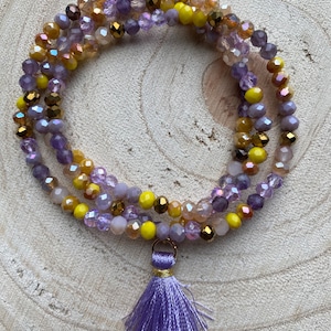 Könnte beinhalten: Ein mehrreihiges Armband mit lila, gelben und goldenen Perlen, abgeschlossen mit einem lila Quaste.