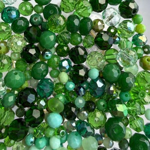 Può includere: Una collezione di perline verdi di varie forme e dimensioni. Le perline variano in tonalità, dal verde menta chiaro al verde smeraldo intenso, con superfici sfaccettate e lisce. Alcune perline sono traslucide, altre opache.