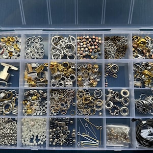 Könnte beinhalten: Ein durchsichtiger Kunststoff-Aufbewahrungsbehälter mit vierundzwanzig Fächern, gefüllt mit verschiedenen Schmuckbastelmaterialien. Das Set enthält Metallringe, Perlen, Verschlüsse, Ohrhaken, Quetschperlen und schwarze Kordel in Gold-, Silber- und Bronzetönen.