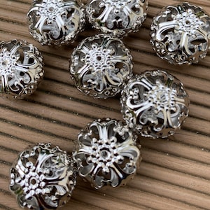 May include: Eight silver metal beads with a decorative, openwork design. The beads have a floral pattern and a raised center.