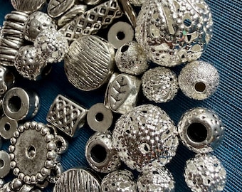 May include: A collection of assorted silver-toned metal beads in various shapes and textures, including spherical filigree beads, flat circular discs, rectangular pieces with grid patterns, and small beads featuring engraved leaf motifs.