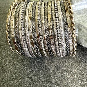 Può includere: Una collezione di braccialetti d'argento e oro. Alcuni braccialetti sono adornati con piccole pietre scintillanti, mentre altri presentano motivi e texture intricati. I braccialetti sono legati con un nastro bianco.
