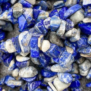 Könnte beinhalten: Nahaufnahme eines Haufens blauer und weißer, getrommelter Lapislazuli-Steine. Die Steine sind unregelmäßig geformt und haben eine natürliche, raue Textur.