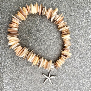 Könnte beinhalten: Ein beige-braunes Muschel-Armband mit einem silbernen Seestern-Anhänger. Das Armband besteht aus vielen kleinen, flachen Muscheln, die übereinander gestapelt sind.