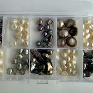 Peut inclure: Une boîte de rangement en plastique transparent dotée de dix compartiments individuels remplis d'un assortiment de perles d'eau douce irrégulières dans des tons variés de blanc, crème, gris, marron et de teintes métalliques sombres.