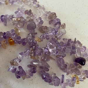 Può includere: Una collana di perline di pietra preziosa di colore viola e oro. Le perline sono di forma e dimensione irregolari e sono infilate su un filo trasparente.