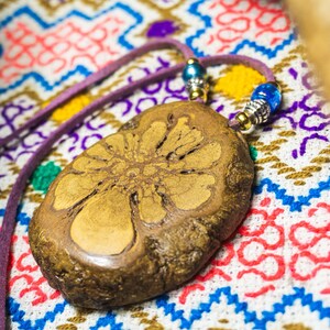 Può includere: Un ciondolo in legno marrone con un design floreale dettagliato, appeso a un cordoncino viola con perline argentate e blu.