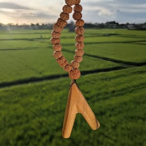 Puede incluir: Un collar que presenta una hilera de cuentas redondas y texturizadas de color marrón que conducen a un colgante de madera lisa de color marrón claro con forma de V. El colgante está unido a las cuentas por un fino cordón negro.