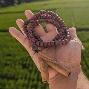 Peut inclure: Un collier composé d'un rang de petites perles en bois rondes et mates de couleur brun-rouge foncé, avec un pendentif en bois lisse en forme de V d'une teinte plus claire, relié par un cordon sombre et une perle foncée à la base.