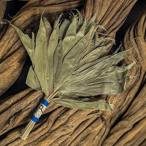 Può includere: Un ventaglio di palma essiccata con un fascio di steli avvolti in tessuto blu e un disegno bianco e nero. Le foglie del ventaglio sono di un colore verde tenue. Il ventaglio poggia su uno sfondo di legno marrone strutturato.