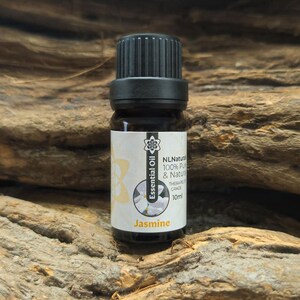 Può includere: Una piccola bottiglia di vetro marrone di olio essenziale di gelsomino con tappo nero. L'etichetta recita "Essential Oil" e "Jasmine", con un disegno floreale. La bottiglia contiene 10 ml di olio.