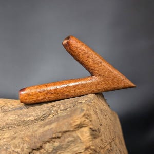 May include: A wooden arrowhead shaped pipe, made of dark brown wood with a reddish-brown hue. The pipe is resting on a piece of rough-hewn wood.