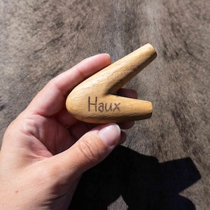 Może przedstawiać: Jasna drewniana fajka z wygrawerowanym słowem "Haux". Fajka ma zakrzywiony kształt z ustnikiem i cybuchem. Widoczne jest usłojenie drewna. Fajka jest trzymana w dłoni.