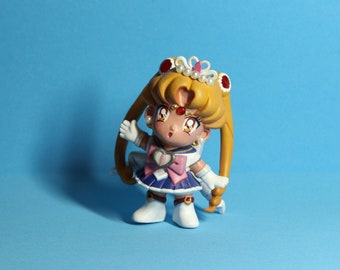 May include: A small, hand-painted figurine of a character with blonde hair styled in two long pigtails. The figure wears a sailor-style outfit with a pink bow, white gloves, and boots. A tiara with pearls and a heart-shaped brooch complete the look.