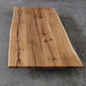 Könnte beinhalten: Eine rechteckige Tischplatte aus Holz mit einer natürlichen Baumkante an einer Seite. Die Oberfläche zeigt eine hellbraune Maserung mit sichtbaren dunklen Ästen, Holzstreifen und organischen Texturen über die verbundenen Holzplanken hinweg.