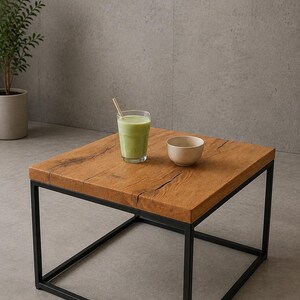 Könnte beinhalten: Ein quadratischer Couchtisch mit einer dicken Tischplatte aus naturfarbenem Holz mit sichtbaren Rissen und einem schwarzen Metallrahmen. Auf dem Tisch stehen ein klares Glas mit einem hellgrünen Smoothie und einem Strohhalm sowie eine kleine, schlichte beige Keramikschale.