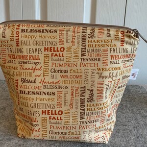 May include: A beige zippered pouch with a brown and orange print of various fall-themed words, including "Welcome Fall", "Happy Harvest", "Fall Greetings", "Thankful", "Pumpkin Patch", "Glorious Fall", and "Blessed Harvest".