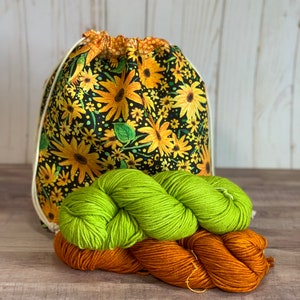 May include: A drawstring bag with a sunflower and floral print design. The bag is filled with two skeins of yarn, one green and one orange.