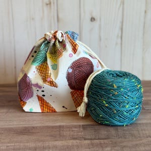 May include: A drawstring project bag with a colorful ice cream cone pattern next to a teal yarn ball. The bag is cream-colored with brown, orange, and purple ice cream cone designs. The yarn ball has a white cord and tassel.