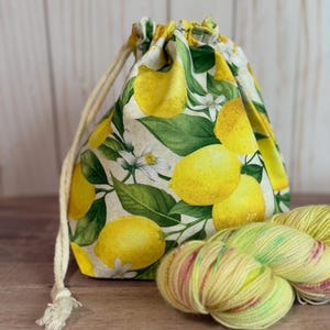 May include: A drawstring project bag with a lemon print, featuring yellow lemons, green leaves, and white blossoms. The bag is next to three skeins of yellow and pink yarn, perfect for knitting or crochet projects. The bag has a beige drawstring.