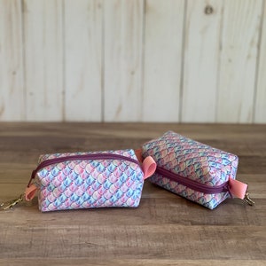 Peut inclure: Deux petites pochettes zippées avec un motif d'écailles de sirène rose, bleu et blanc. Les pochettes ont une fermeture éclair violette et une sangle rose.