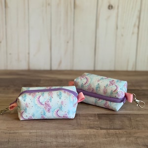 Peut inclure: Deux petites pochettes zippées avec un fond bleu clair et un motif floral violet et rose. Les pochettes ont des tirettes de fermeture éclair roses et une fermeture éclair violette. Une pochette est munie d'un porte-clés argenté.