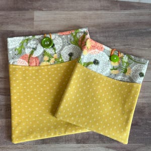 May include: Two fabric book sleeves. The top section has a grey background with white dandelions and peach flowers. The bottom is yellow with a white diamond pattern. Each sleeve has a green button closure.