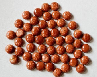 May include: A collection of round, polished, golden-brown gemstones with a sparkling, glittery appearance. The gemstones are arranged closely together on a light-colored surface, showcasing their smooth, domed shape and reflective qualities. These stones are ideal for jewelry making.