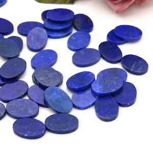 Puede incluir: Una colección de cabujones de lapislázuli ovalados, cada uno con un color azul intenso y motas de pirita dorada. Las piedras están esparcidas sobre una superficie blanca.