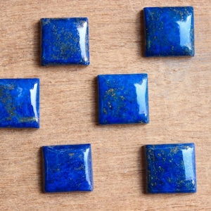 Peut inclure: Six cabochons carrés en lapis-lazuli avec des inclusions dorées sur une surface en bois.