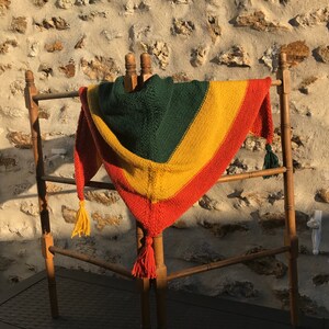 May include: A colourful knitted shawl, triangular in shape, draped over a wooden clothes horse. The shawl is striped in green, yellow, and orange, with tassels at the points. The background is a stone wall.