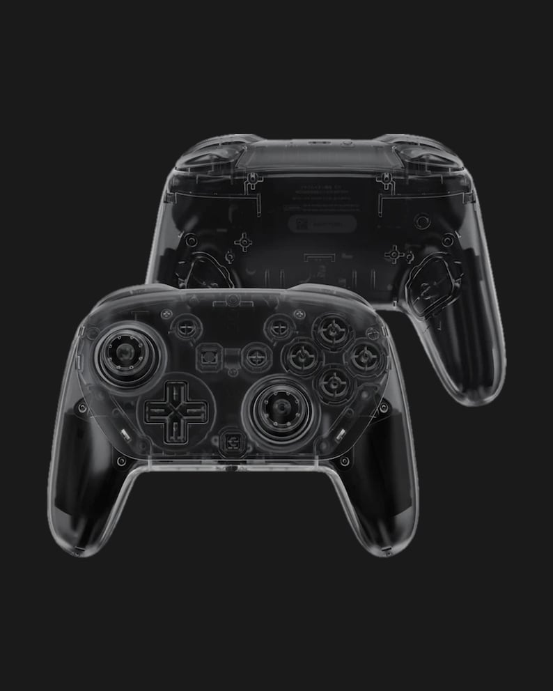May include: Two transparent game controllers are displayed against a black background. The controllers have a clear plastic shell, revealing the internal components. The design includes buttons, joysticks, and a directional pad, typical of modern game controllers.
