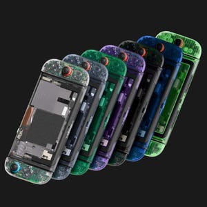 May include: A series of seven handheld gaming consoles arranged in a diagonal row. Each device features a transparent plastic shell in various colors including clear, grey, green, purple, black, and blue, revealing internal components.