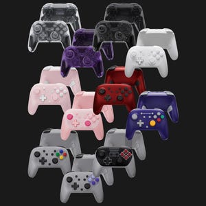 May include: A collection of various game controllers in a variety of colors, including clear, black, white, pink, red, and blue. The controllers have buttons, joysticks, and directional pads.