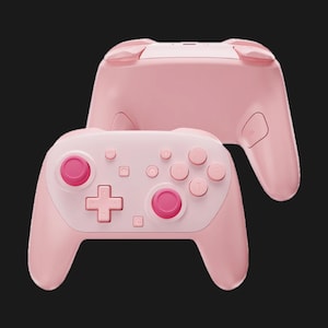 May include: Two pink game controllers are displayed against a black background. The controllers have a light pink body with white and pink buttons. The buttons include a directional pad, face buttons, and analog sticks. One controller is in front, and the other is behind it.
