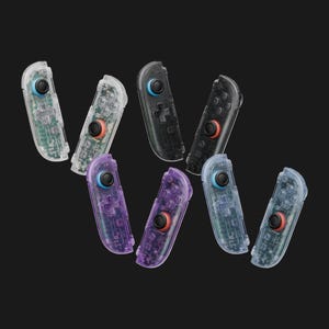 May include: A collection of eight gaming controllers featuring transparent shells in clear, black, and purple hues. Each controller includes a circular joystick with either a blue or red cap, a directional pad, and four action buttons visible through the casing.