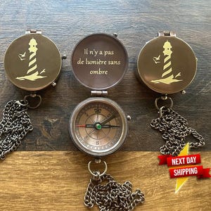 Peut inclure: Un compas en laiton avec une chaîne, représentant un phare et la citation "Il n'y a pas de lumière sans ombre".