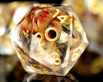 May include: A clear, multi-sided dice with gold numbers and a red and yellow flower pressed inside. The dice is sitting on a black surface.