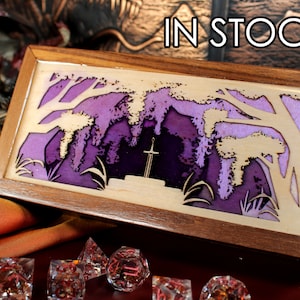 May include: A wooden box with a purple and white stained glass design of a forest scene with a sword in the centre. The box is in stock.