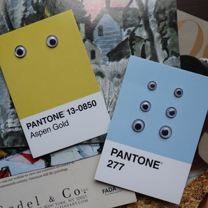 Può includere: Due schede colore Pantone, Aspen Gold e 277, ciascuna con occhi mobili attaccati, poggiano su una pila di riviste e materiali stampati. La scheda Aspen Gold ha due occhi, mentre la 277 ne ha sei. Le schede sono un'interpretazione giocosa delle mazzette colori.